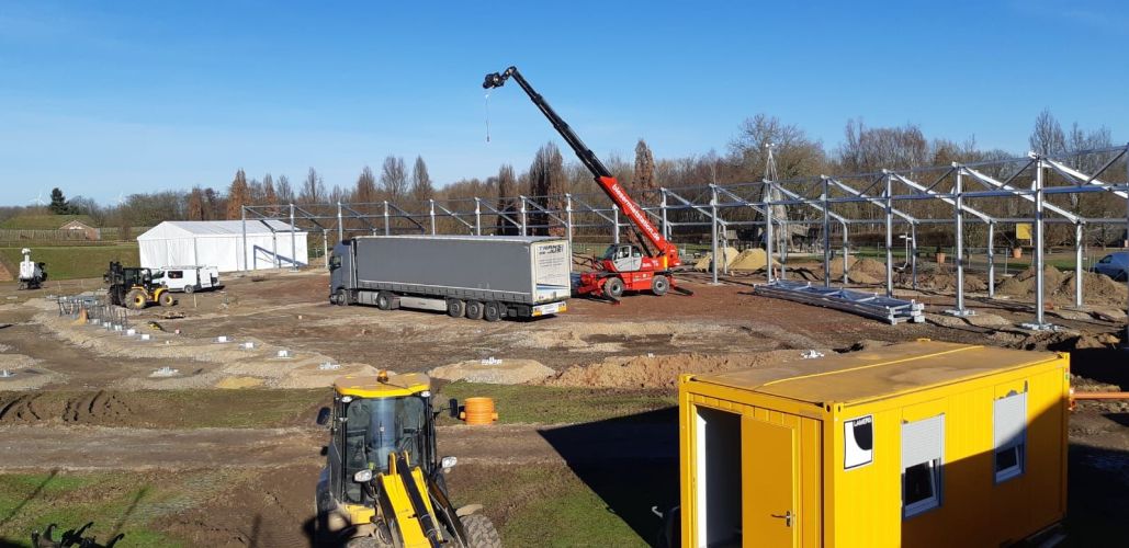 Bild einer Baustelle, auf der gerade eine Halle errichtet wird. Es sind nur ein Krahn und erste Elemente des Grundgerüsts der Halle sichtbar